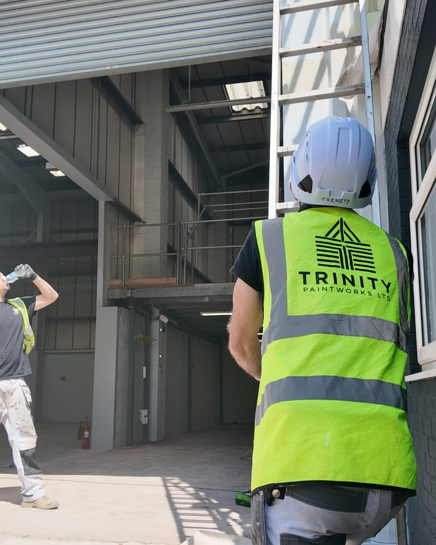 A Trinity Paintworks painter in a Trinity hi-vis vest and hard hat footing a ladder outside an industrial unit